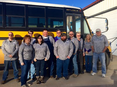 Photo of all the Bus Driver of the school, standing before a row of yellow buses and smiling at the camera.