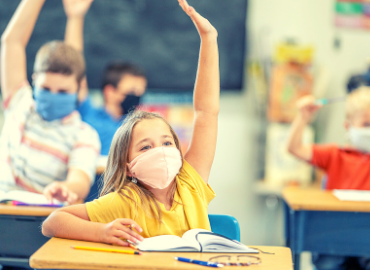 Girl in a yellow shirt, wearing a mask, raising her hand in a classroom