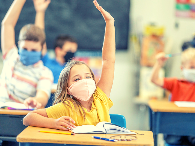 little girl in yellow shirt, wearing a mask, raising her hand in a classroom
