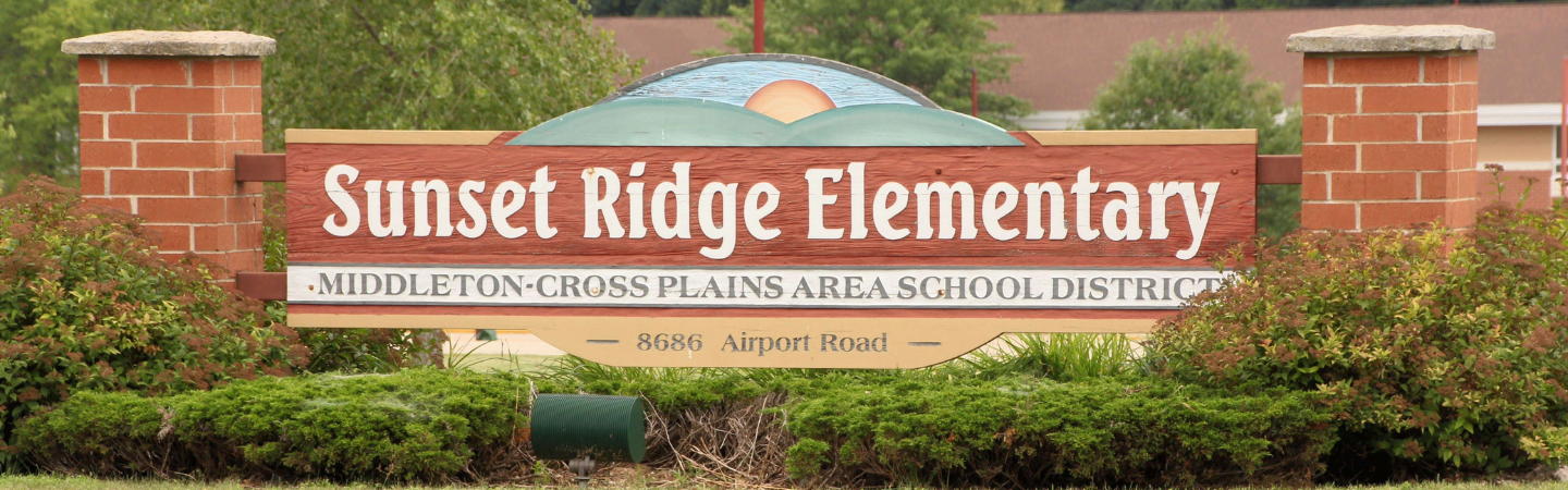 Picture of the sign "Sunset Ridge Elementary" school sign. The sign has an image painted on it depicting green hillsides with the sun rising against a blue sky. The school address, 8686 Airport Road is included on the sign.